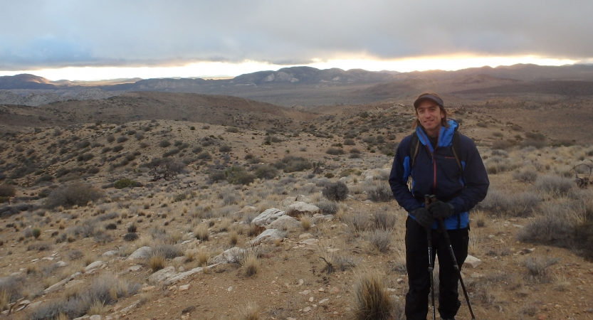 A person wearing a backpack and holding a trekking pole smiles at the camera amidst the desert landscape of Joshua Tree National Park.